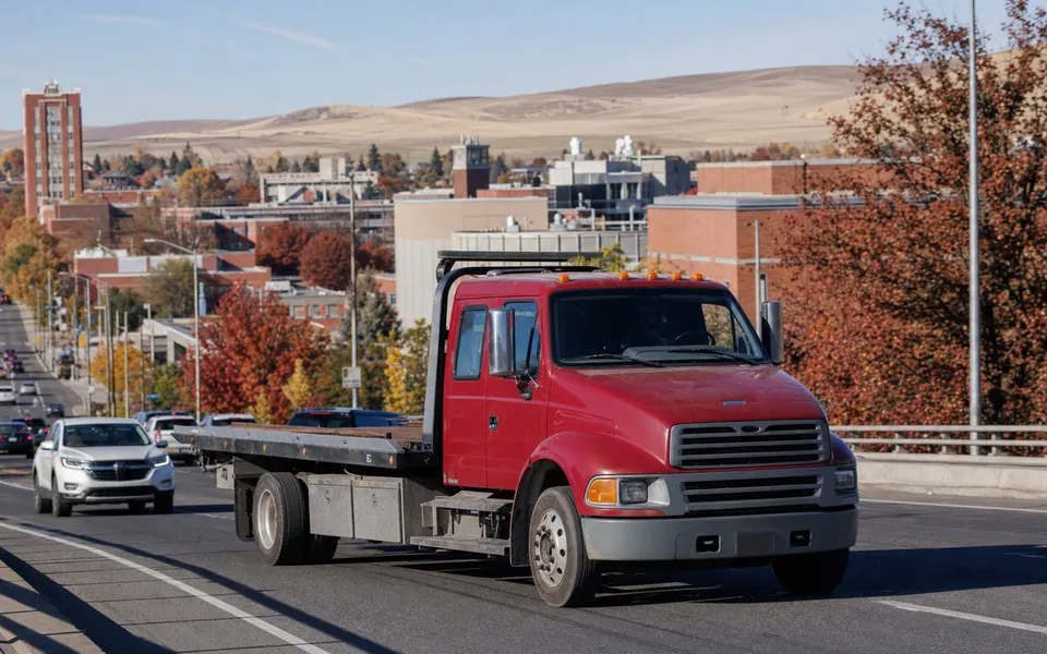 Crimson flatbed tow truck traveling through Pullman with campus-area buildings and Palouse hills in the background