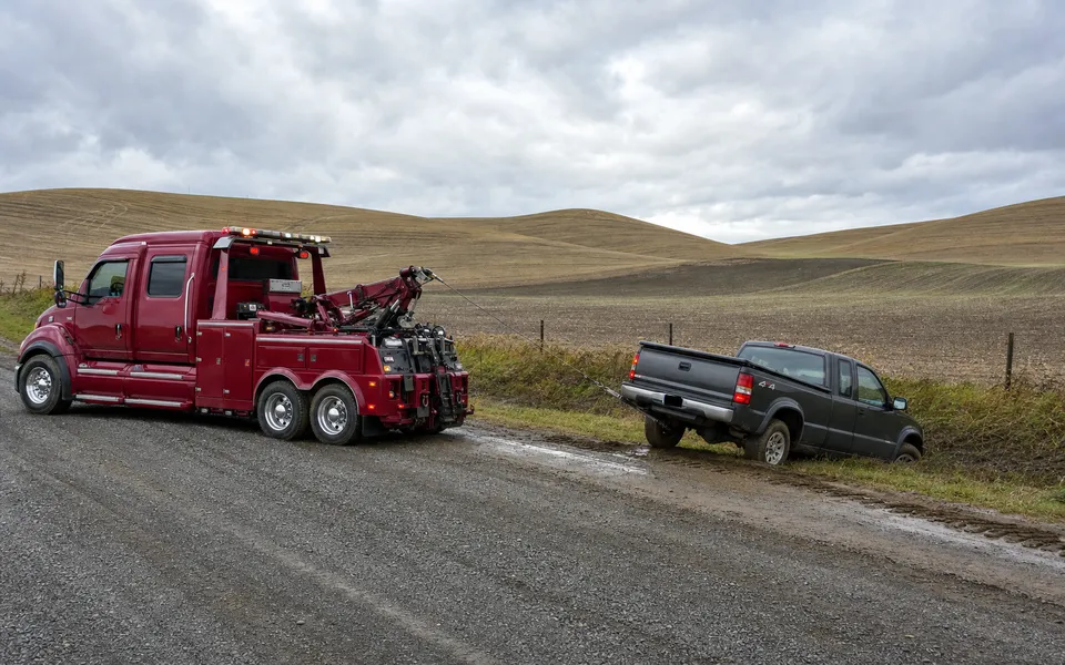 Crimson tow truck winching a pickup from a muddy gravel shoulder among the rolling Palouse hills near Pullman, Washington